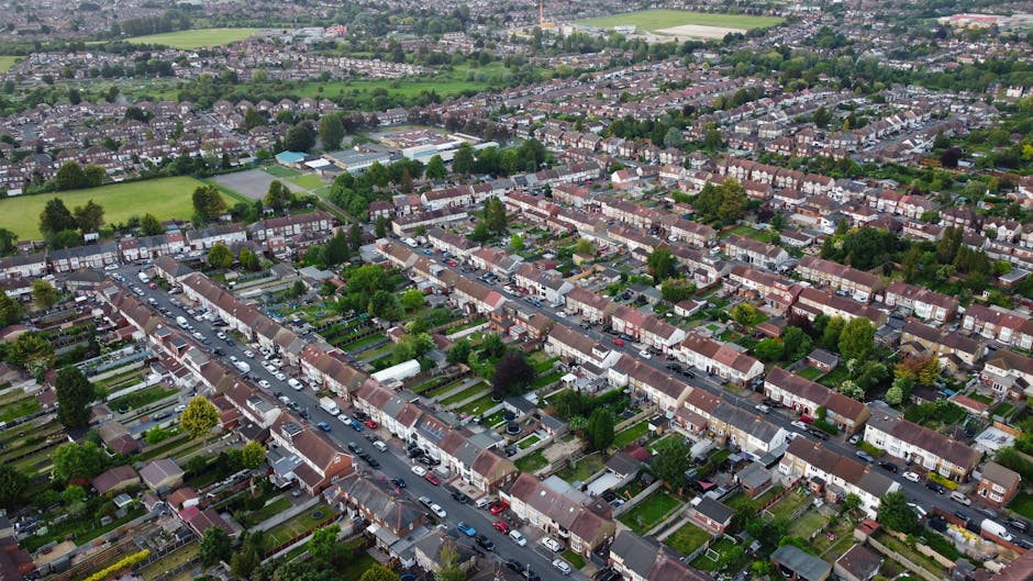 Aerial view of a residential suburb showing rows of terraced houses with small front gardens and driveways, some with cars parked on them or along the street. The houses have brick facades with tiled roofs, and many have recycling bins or garden furniture visible in the yards. A narrow road runs through the centre of the image with parked vehicles on both sides, leading towards larger green spaces and sports fields in the distance. The scene captures a typical UK neighbourhood environment suitable for house removals and furniture transport logistics, as part of a home relocation process. The natural lighting indicates daytime, with trees and greenery interspersed among the houses, providing a quiet and organized community setting, relevant to moving and packing services offered by Man with Van Orpington during local house removals.