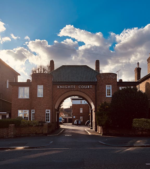 A photograph of the entrance to Knights Court, a residential building complex with a brick archway and two surrounding brick buildings, taken during daylight hours with a partly cloudy sky overhead. The archway has the words 'KNIGHTS COURT' displayed above it, and through the archway, a paved driveway and parked cars are visible. To the right of the entrance, a large bush and some greenery are present, with the building's chimneys visible against the sky. The image captures the exterior environment of a typical home relocation setting, with the building's facade and external features clearly visible, supporting landscape and property context for removals and transport services.