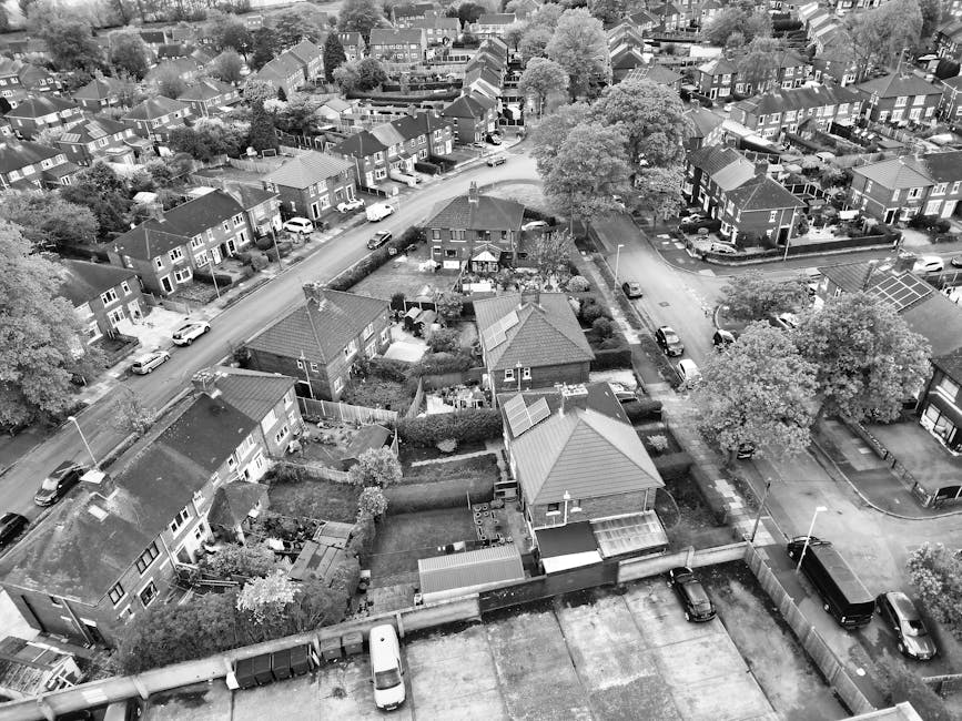 Aerial view of a residential suburb showing rows of terraced houses with small front gardens and driveways, some with cars parked on them or along the street. The houses have brick facades with tiled roofs, and many have recycling bins or garden furniture visible in the yards. A narrow road runs through the centre of the image with parked vehicles on both sides, leading towards larger green spaces and sports fields in the distance. The scene captures a typical UK neighbourhood environment suitable for house removals and furniture transport logistics, as part of a home relocation process. The natural lighting indicates daytime, with trees and greenery interspersed among the houses, providing a quiet and organized community setting, relevant to moving and packing services offered by Man with Van Orpington during local house removals.