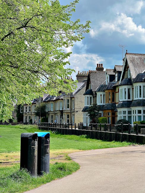 The image shows a row of traditional detached houses with bay windows, pitched roofs, and chimneys, situated behind a black metal fence on a residential street. In the foreground, there are two black wheelie bins placed on a grass verge adjacent to a concrete pathway. A large tree with green leaves extends across the top left corner of the image, partially covering the sky, which features some scattered clouds. The scene is illuminated by daylight, indicating a bright, sunny day. The environment appears well-maintained, and the setting is typical of a suburban area suitable for home relocations or moving services. This image aligns with the context of house removals and furniture transport, illustrating a typical residential street where relocation activities may take place, as highlighted in the 'Bulky Waste Removal Costs in Orpington' page on manwithvanorpington.co.uk.