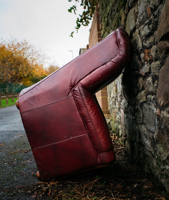 A red leather armchair has been dislodged from a property and is now positioned partially on its side, leaning against a stone wall on a paved outdoor pathway. The chair’s backrest and armrests are visible, with the furniture showing some signs of wear and creasing on the leather upholstery. Surrounding the chair, there are scattered leaves and debris, indicating an outdoor setting. In the background, a garden area and a section of a brick house are visible, with some trees displaying autumn foliage under an overcast sky. This scene captures the process of furniture removal during a home relocation or moving service, possibly part of a larger move handled by Man with Van Orpington, who specialise in removals and waste clearances. The arrangement suggests the chair is being loaded onto a vehicle, such as a van, for transport to a new location, in line with packing and moving logistics.