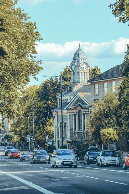 A street scene in Orpington featuring a large Victorian-style church with a tall clock tower topped by a spire, situated behind a row of parked cars along the pavement. The church's exterior is made of light-colored brick with decorative elements, and its windows are arched. To the left, there are leafy green trees and modern streetlights, while the sky above is partly cloudy with patches of blue. The street appears calm with minimal traffic, reflecting typical conditions during a house removal or relocation process. Although no furniture, boxes, or moving equipment are visible in this image, it provides context for a residential area where professional removals, such as those offered by Man with Van Orpington, would facilitate home relocation, furniture transport, and packing and moving services in the Crofton Road area.