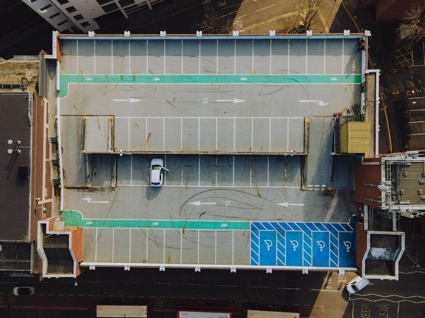 An aerial view of a building rooftop parking area showing designated parking bays, including specific blue disabled parking spaces at the bottom right corner. The area is primarily paved with concrete