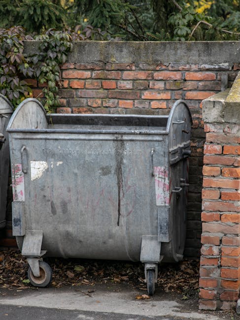 A large, grey metal waste collection skip positioned outdoors against a brick wall with some green leafy plants growing above it. The skip has visible signs of weathering and rust, with pink and white markings faintly visible on its surface. It is mounted on four small wheels, one of which is partially buried in dirt and leaves on the ground, indicating it is stationary. The skip is located on a paved area adjacent to a door or entrance, suggesting its use for waste disposal during home relocation or renovation activities. The environment appears to be a residential or service area, with natural daylight illuminating the scene. This image reflects the typical waste management setup associated with moving or clearing a property, as handled by companies like Man with Van Orpington, which often coordinate the removal of waste and furniture transport during house removals and logistics.