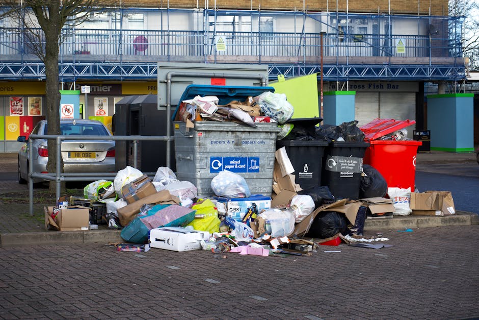A collection of overflowing public waste and recycling bins situated on a paved sidewalk in front of a retail building with a yellow and black sign. The waste bins include a large grey mixed paper and card container, a black general waste bin, and a red recycling bin, all filled to capacity with various cardboard boxes, paper, plastic packaging, and black rubbish bags spilling onto the ground. There are additional discarded packaging materials, small cardboard boxes, and plastic bags scattered around the bins. A metal railing separates the bins from parked cars on the street. Behind the bins, a multi-storey building with scaffolding and protective netting is visible, indicating construction or maintenance work. The scene is lit by natural daylight, and the area appears to be a designated collection point for household waste, relevant to topics such as house removal and waste disposal logistics, as handled by companies like Man with Van Orpington.