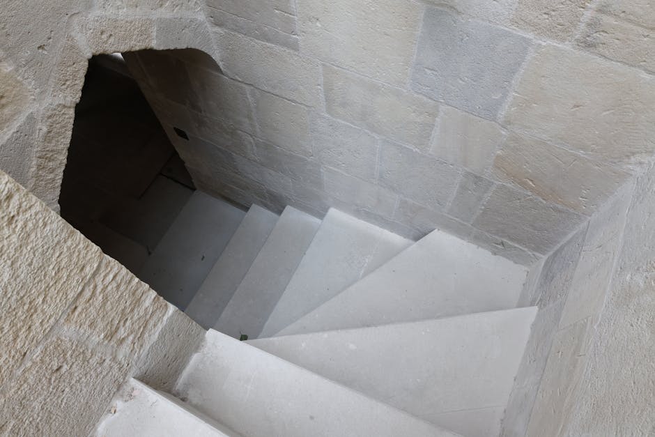 Looking down a narrow, inward-facing staircase made of light-colored stone, with seven visible steps descending into a dark basement or lower level. The staircase is enclosed by stone walls on both sides, with rough textured bricks on the lower part and smoother stone blocks higher up. At the top of the stairs, there is an arched opening leading into the space, and the surrounding area indicates an indoor residential setting, possibly during a home relocation process. The lighting appears natural, illuminating the treads and the stonework, while the shadows accentuate the depth of the staircase. This image exemplifies the type of challenging access that Man with Van Orpington can assist with during house removals or furniture transport, especially when navigating narrow staircases.