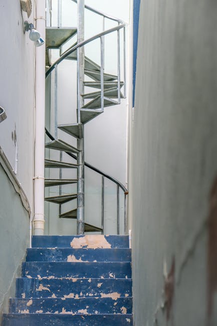 A metal spiral staircase with a rounded black handrail ascending inside a narrow, well-lit interior space. The blue-painted concrete stairs at the base show signs of wear with chipped paint and scuffs. To the left, there's a white pipe running vertically along the wall, and part of a white door is visible. The surrounding walls are plain and painted in neutral tones, with some damage or peeling near the bottom. This staircase is part of a residential property, and the image captures the location where [COMPANY_NAME] might be involved in home relocation or furniture transport, preparing for moving or packing processes within the property interior.