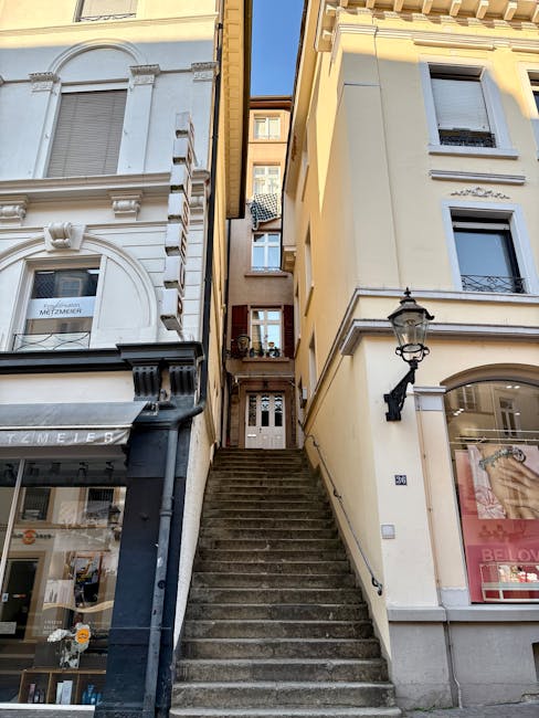 Looking down a narrow, inward-facing staircase made of light-colored stone, with seven visible steps descending into a dark basement or lower level. The staircase is enclosed by stone walls on both sides, with rough textured bricks on the lower part and smoother stone blocks higher up. At the top of the stairs, there is an arched opening leading into the space, and the surrounding area indicates an indoor residential setting, possibly during a home relocation process. The lighting appears natural, illuminating the treads and the stonework, while the shadows accentuate the depth of the staircase. This image exemplifies the type of challenging access that Man with Van Orpington can assist with during house removals or furniture transport, especially when navigating narrow staircases.