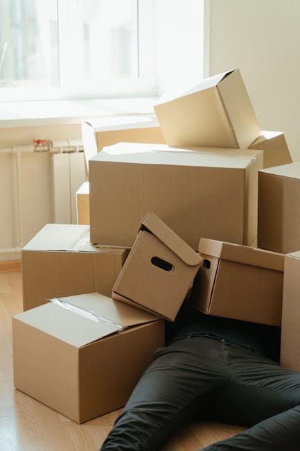 A person wearing black trousers lying on the floor inside a room, partially obscured by multiple stacked cardboard moving boxes of various sizes, some sealed with packing tape and others open or partially unpacked. The boxes are placed near a window with natural light streaming in, highlighting the contents and the room's light-colored walls. The scene depicts the packing or unpacking stage of a house move, with a focus on the logistics of furniture transport and packing materials, consistent with services offered by Man with Van Orpington. The environment appears tidy, and the presence of boxes suggests ongoing home relocation preparations or post-move organization.
