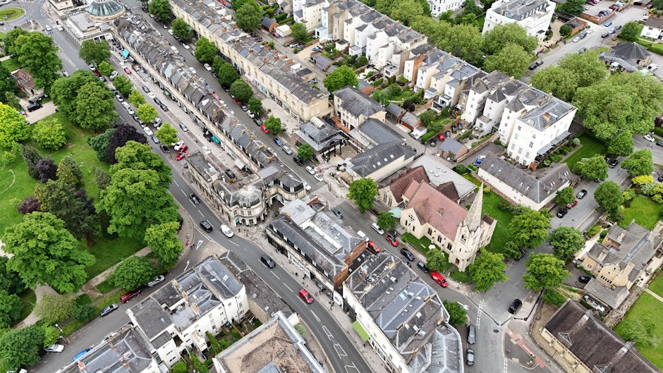 An aerial view of a residential neighbourhood in Orpington, showing a narrow street lined with parked cars on both sides, surrounded by a mix of terraced and semi-detached houses with grey and brown roofs. In the foreground, a small church with a steeple and beige walls is visible beside the street, with green trees and grassy areas nearby. The street appears to be part of a home relocation or moving process, consistent with the services offered by Man with Van Orpington, including furniture transport and packing during house removals.