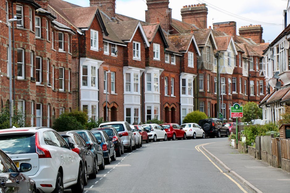A residential street in Orpington featuring a row of traditional red-brick terraced houses with bay windows and pitched roofs. Several parked cars, including sedans and hatchbacks, line both sides of the narrow, curved pavement, with some vehicles partially obstructing the sidewalk. A green and red sign indicating a residents' parking zone is visible near the curb. The street is lit by a lamppost, and the sky is partly cloudy, providing natural daylight. The image depicts a typical setting where home relocation or furniture transport might involve careful parking and driveway access, emphasizing space constraints common to house removals in tight driveways. Man with Van Orpington specializes in removals and household moving services, often navigating such urban environments during the loading process or home packing and moving activities.
