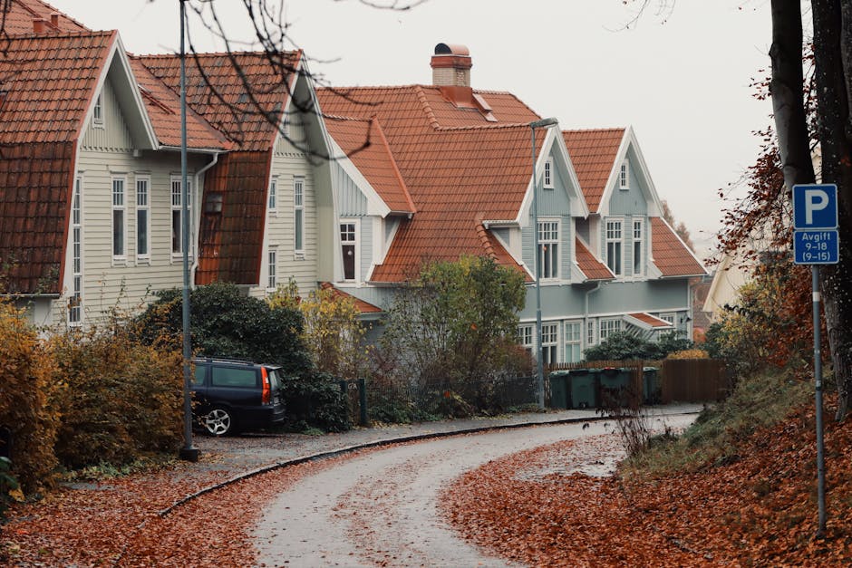 A narrow, curved driveway in a residential area with a gravel surface, bordered by autumnal trees and bushes with orange and brown leaves. The driveway leads to a row of traditional-style houses with white wooden exteriors and red-tiled, gabled roofs. A black car is parked near the front of one house, partially obscured by shrubs. A blue parking sign indicating parking restrictions is mounted on a pole on the right side of the driveway. The scene is overcast with diffused natural light, and the overall setting suggests a quiet neighborhood suitable for home relocation or furniture transport by a professional moving service such as Man with Van Orpington, who may assist with house removals in areas with limited driveway space.